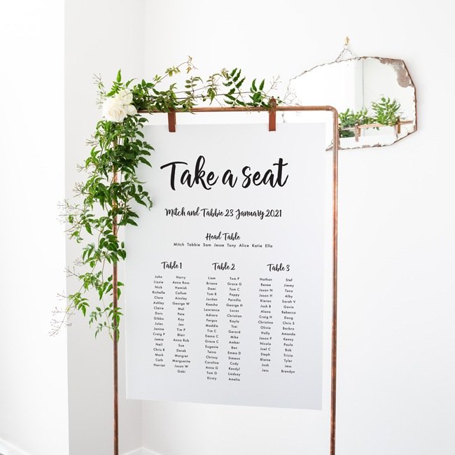 large seating plan hanging from metal frame with flowers that says take a seat